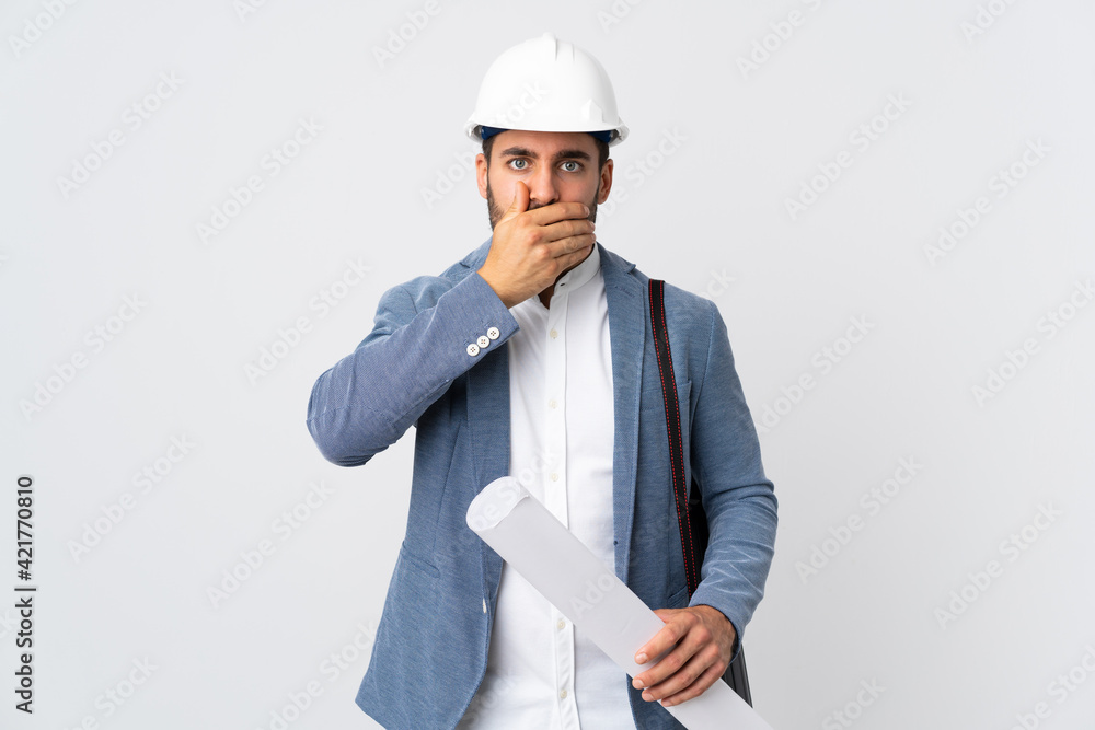 Young architect man with helmet and holding blueprints isolated on white background covering mouth with hand Young architect man with helmet and holding blueprints isolated on white background covering mouth with hand