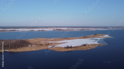 Aerial waterscape with a small island on a river with parly frozen water and overgrown with dry reeds and trees at sunny spring day photo