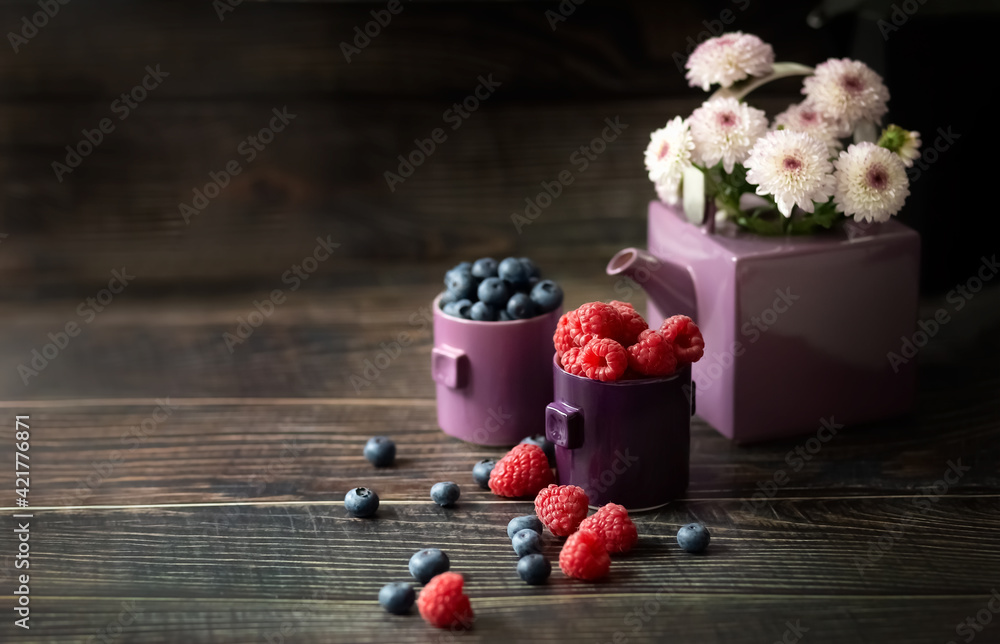 Berries raspberries and blueberries in a cup on a wooden table. Chrysanthemums in the teapot. Still life. Copy space. Place for your text.