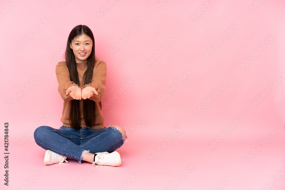 Young asian woman sitting on the floor isolated on pink background holding copyspace imaginary on the palm to insert an ad Young asian woman sitting on the floor isolated on pink background holding copyspace imaginary on the palm to insert an ad