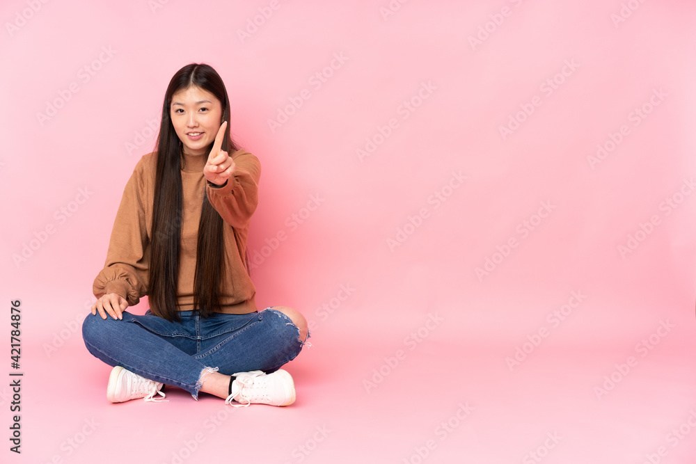 Young asian woman sitting on the floor isolated on pink background showing and lifting a finger Young asian woman sitting on the floor isolated on pink background showing and lifting a finger