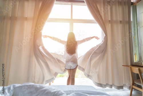 Young asian woman stand open white curtains at the window, the morning after waking up in the bedroom of her home. Woman wake up with a fresh and open the curtains on the windows.