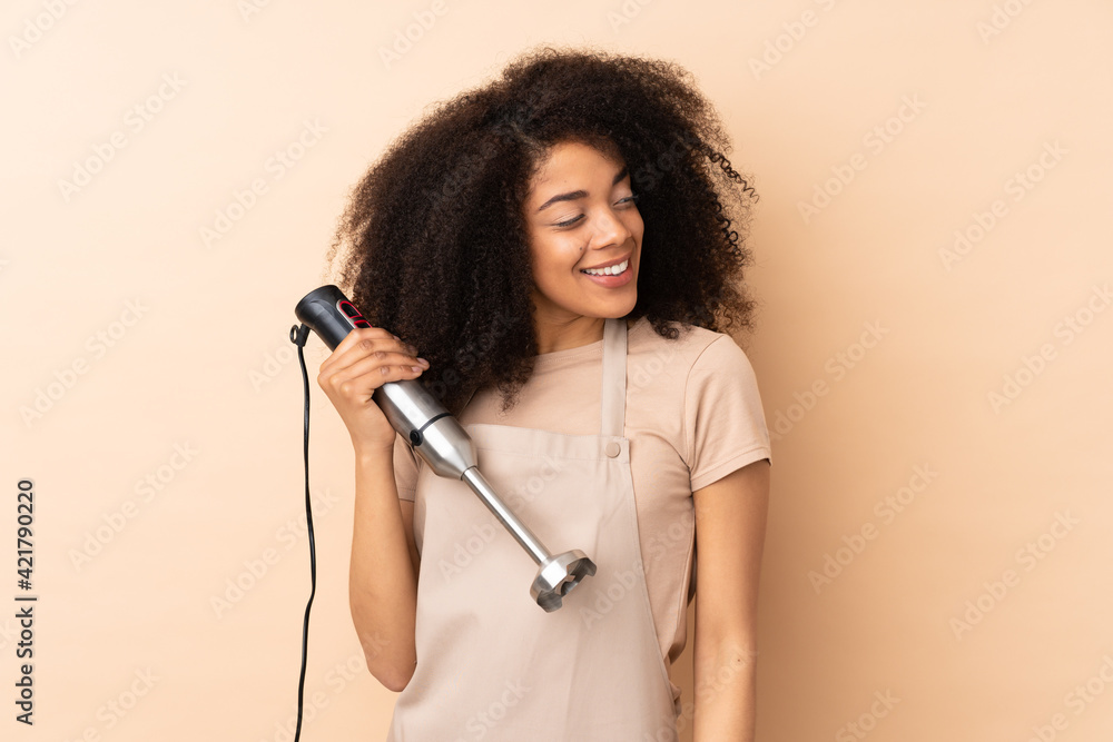 Young african american woman using hand blender isolated on beige background looking to the side Young african american woman using hand blender isolated on beige background looking to the side