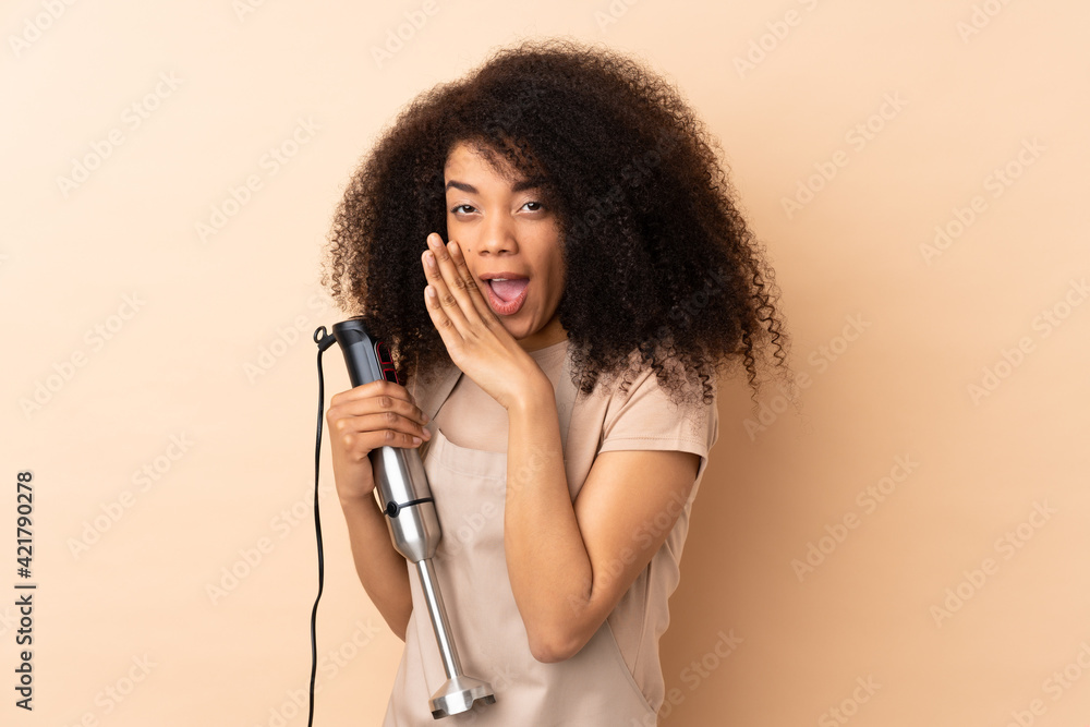 Young african american woman using hand blender isolated on beige background whispering something