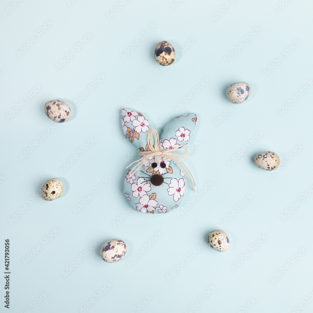 Flat lay Easter symbols: quail eggs laid out in circle around one DIY textile Easter bunny on light blue.