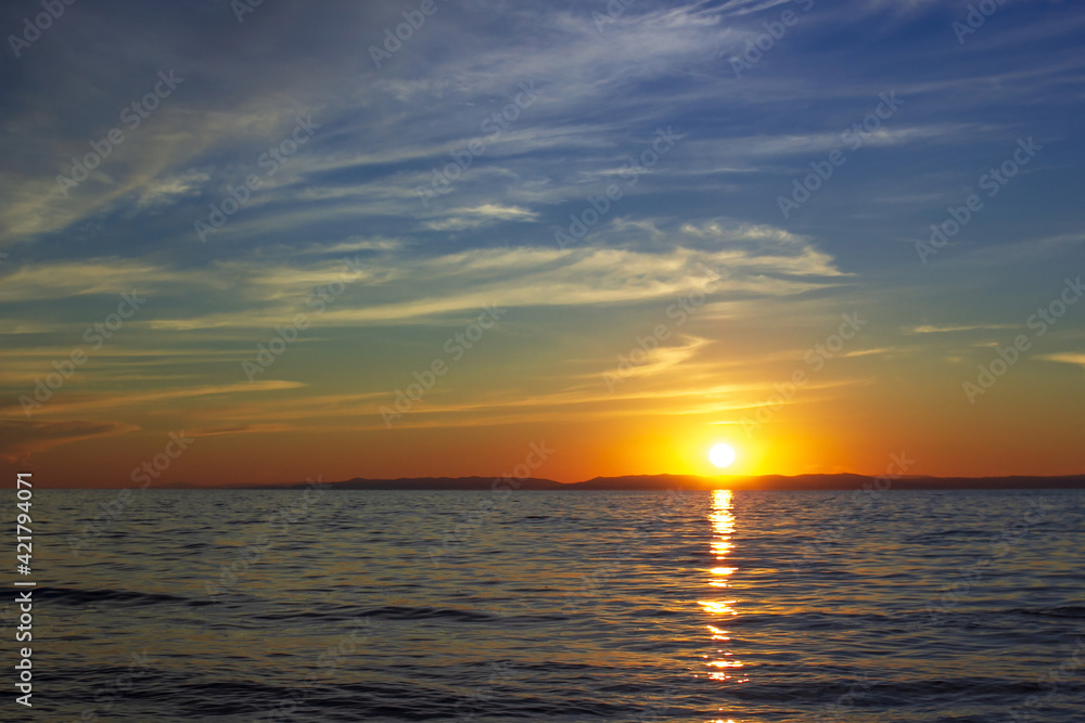 Scenic sunset on Lake Baikal in dark moody colors with blue sky, the bright sun sets over the horizon above calm waves Scenic sunset on Lake Baikal in dark moody colors with blue sky, the bright sun sets over the horizon above calm waves