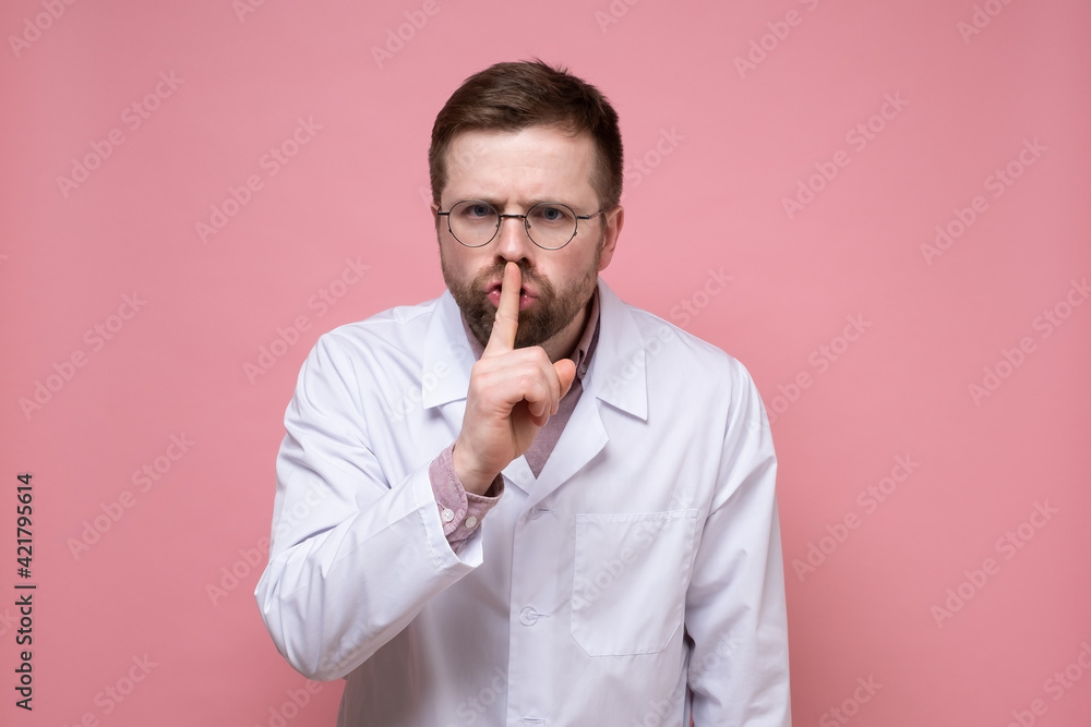 Male doctor with a stethoscope around neck put index finger to lips, making a gesture of silence and looking seriously. Male doctor with a stethoscope around neck put index finger to lips, making a gesture of silence and looking seriously.