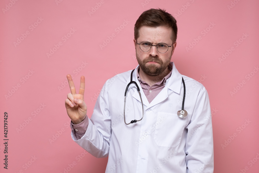 Frustrated male doctor is offended at something, but shows a symbol of victory and frowns. Two fingers gesture. Frustrated male doctor is offended at something, but shows a symbol of victory and frowns. Two fingers gesture.