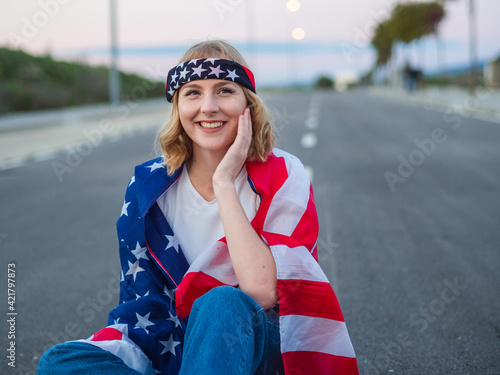 Mujer joven rubia con ojos azules sentada en una carretera celebrando el dia de la independencia de estados unidos photo