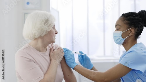 An elderly woman getting a vaccine for coronavirus. Female doctor wearing mask and gloves with a syringe makes an injection to a mature senior patient. Concept of health care, vaccination program. photo