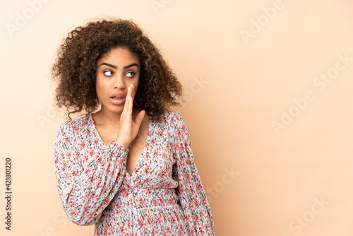 Young African American woman isolated on beige background whispering something with surprise gesture while looking to the side