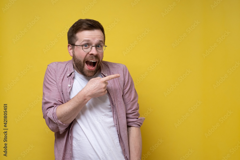Surprised, charming man in a pink shirt and glasses points finger to the side and happily looks at the camera. Copy space. Surprised, charming man in a pink shirt and glasses points finger to the side and happily looks at the camera. Copy space.
