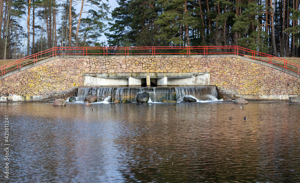 Small waterfall in the city park. Jets of water fall on stones, and ducks swim nearby on the water. Above the waterfall there is a wall of natural stone and a staircase with red railings.
