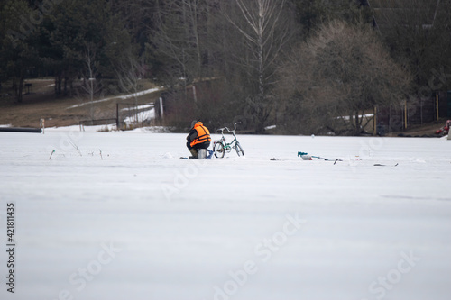 A lone fisherman in a bright life jacket sits on a box on thin spring ice and fishing. Nearby there is a bicycle and an ice screw.