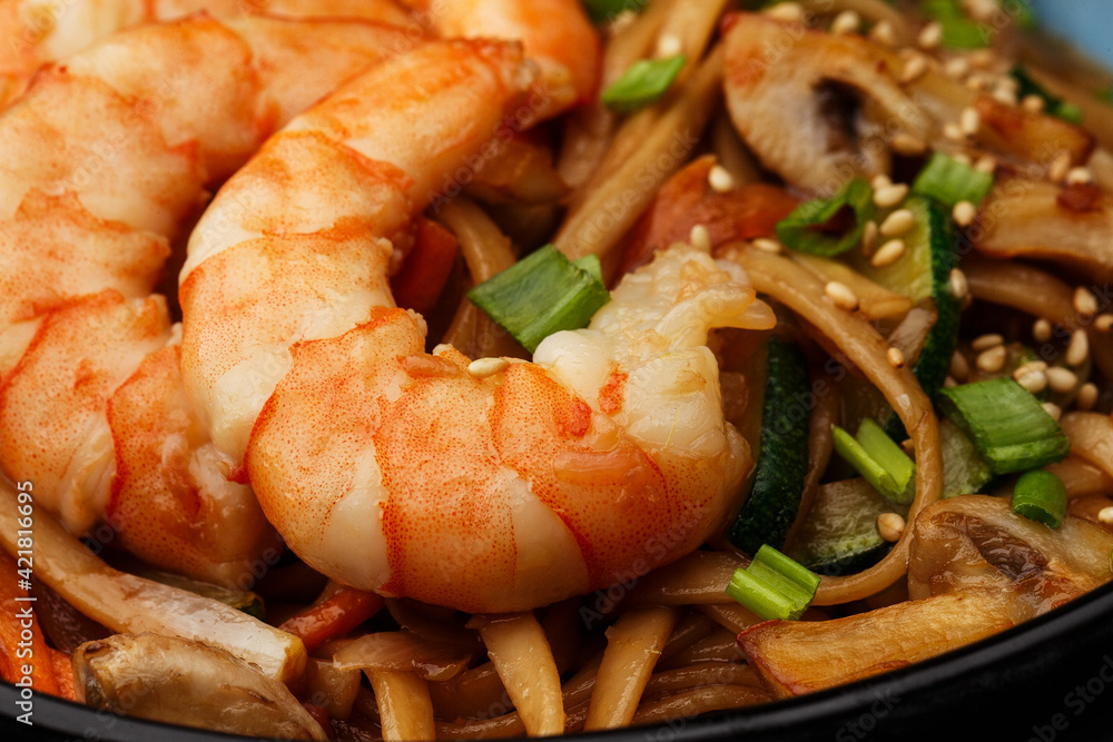 Udon noodles with shrimps and vegetables: zucchini, red pepper, mushrooms, carrot, spring onion and sesame seeds. Dish isolated in a blue bowl, close-up on a black marble background. Asian cuisine. Udon noodles with shrimps and vegetables: zucchini, red pepper, mushrooms, carrot, spring onion and sesame seeds. Dish isolated in a blue bowl, close-up on a black marble background. Asian cuisine.