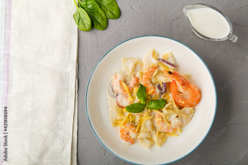 Farfalle pasta with shrimps in a creamy sauce on a gray plate on a concrete background next to a napkin and a jug of cream. Horizontal photo