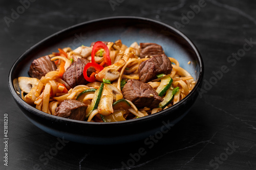 Udon noodles with teriyaki beef and vegetables: zucchini, red pepper, mushrooms, carrot, onion and sesame seeds. Dish isolated in a blue bowl, close-up on a black marble background. Asian cuisine.