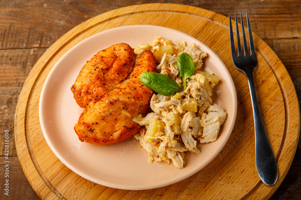 Baked chicken breast in sauce with salad on a plate next to a fork on a wooden table on a round stand. Baked chicken breast in sauce with salad on a plate next to a fork on a wooden table on a round stand.