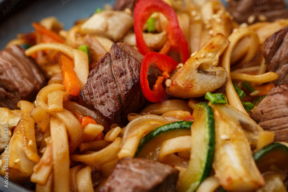 Udon noodles with teriyaki beef and vegetables: zucchini, red pepper, mushrooms, carrot, onion and sesame seeds. Dish isolated in a blue bowl, close-up on a black marble background. Asian cuisine. Udon noodles with teriyaki beef and vegetables: zucchini, red pepper, mushrooms, carrot, onion and sesame seeds. Dish isolated in a blue bowl, close-up on a black marble background. Asian cuisine.