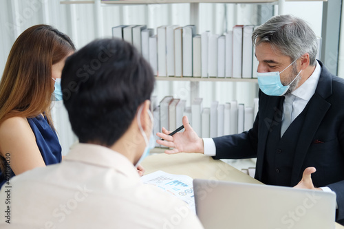 Smart Caucasian senior manager and businessman in black suit talking with asian teamwork staff is wearing masks prevent covid 19 virus at office. New nomal and social distace of business concept. photo