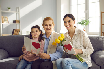 Family portrait of an older woman, her daughter and granddaughter sitting on a sofa and exchanging gifts in honor of Mother's Day. Family holds tulips, handmade cards and looks into the camera.