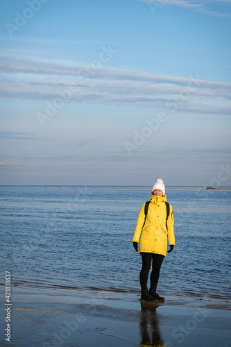 A woman in a yellow winter jacket and with a black backpack walking by the shores of the Baltic Sea near Carnikava, Latvia