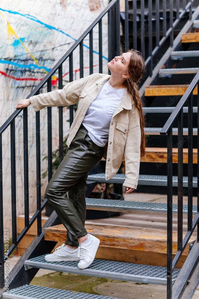 Pretty hipster girl is standing on stairs, Happy beautiful young woman in leather green pants and light beige jacket, urban background, street photo, white sneakers, looking away, casual clothes Pretty hipster girl is standing on stairs, Happy beautiful young woman in leather green pants and light beige jacket, urban background, street photo, white sneakers, looking away, casual clothes
