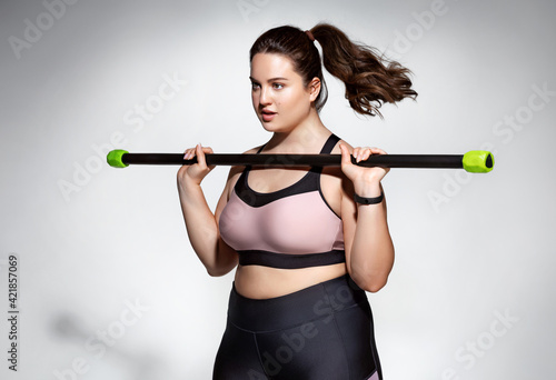 Sporty girl doing exercises with body bar. Photo of model with curvy figure in fashionable sportswear on grey background. Sports motivation and healthy lifestyle