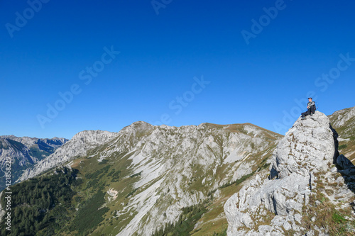 A man with a hiking backpack sitting on the of a big boulder on the way to Hohe Weichsel in Austria, with a panoramic view on a vast valley. Narrow pathway. He is enjoying the view. Discovering