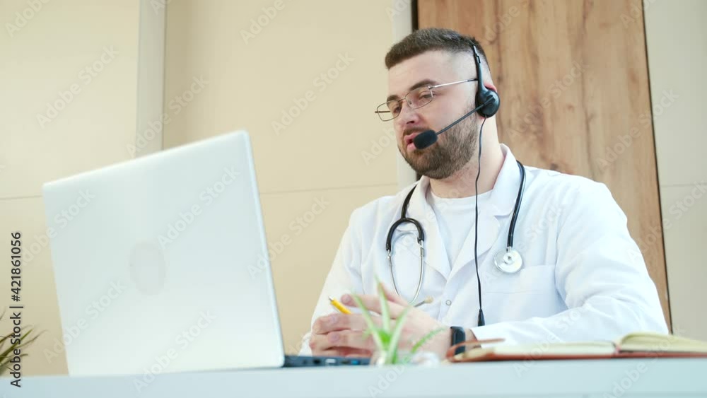 Man professional doctor in white medical coat and headset making conference call on laptop computer, consulting distance patient online in video chat, male explain treatment by webcam. Telemedicine Man professional doctor in white medical coat and headset making conference call on laptop computer, consulting distance patient online in video chat, male explain treatment by webcam. Telemedicine