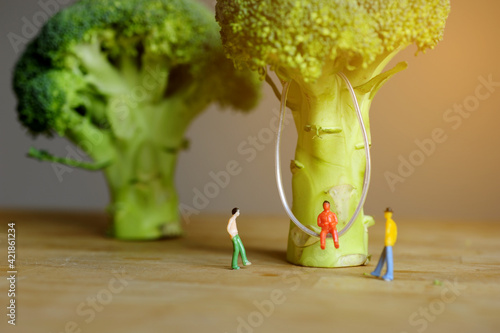 Miniature figurine character: One people sitting on swing that hanging on broccoli and two people standing on wooden floor near by for chatting 