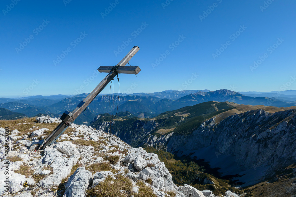 A wooden cross on top of Hohe Weichsel, Alpine peak in Austria. The cross is leaning, as if it was going to fall. There are endless mountain chains behind it. Early fall. The slopes are turning golden A wooden cross on top of Hohe Weichsel, Alpine peak in Austria. The cross is leaning, as if it was going to fall. There are endless mountain chains behind it. Early fall. The slopes are turning golden