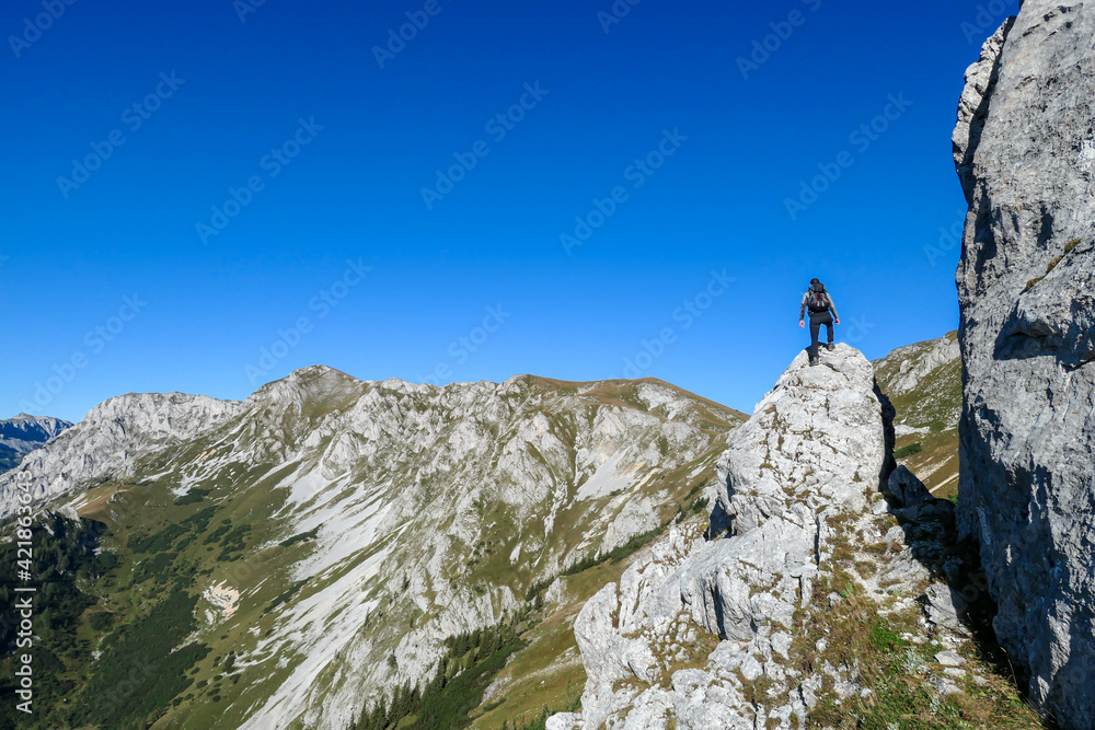 A man with a hiking backpack climbing to the of a big boulder on the way to Hohe Weichsel in Austria, with a panoramic view on a vast valley.  Narrow pathway. He is enjoying the view. Discovering