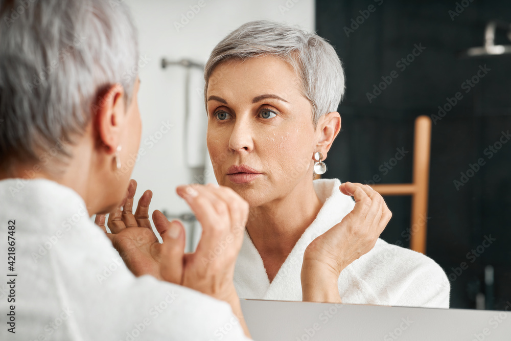 Senior woman with moisturizer on her face looking at mirror in bathroom Senior woman with moisturizer on her face looking at mirror in bathroom