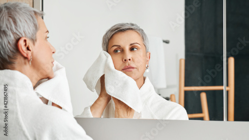 Beautiful mature woman doing morning routine in bathroom wiping the face