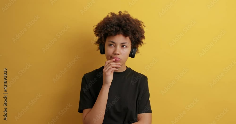 Pensive curly haired woman looks around with thoughtful expression makes assumptions needs to think listens music via wireless headphones dressed in casual black t shirt isolated on yellow background Pensive curly haired woman looks around with thoughtful expression makes assumptions needs to think listens music via wireless headphones dressed in casual black t shirt isolated on yellow background