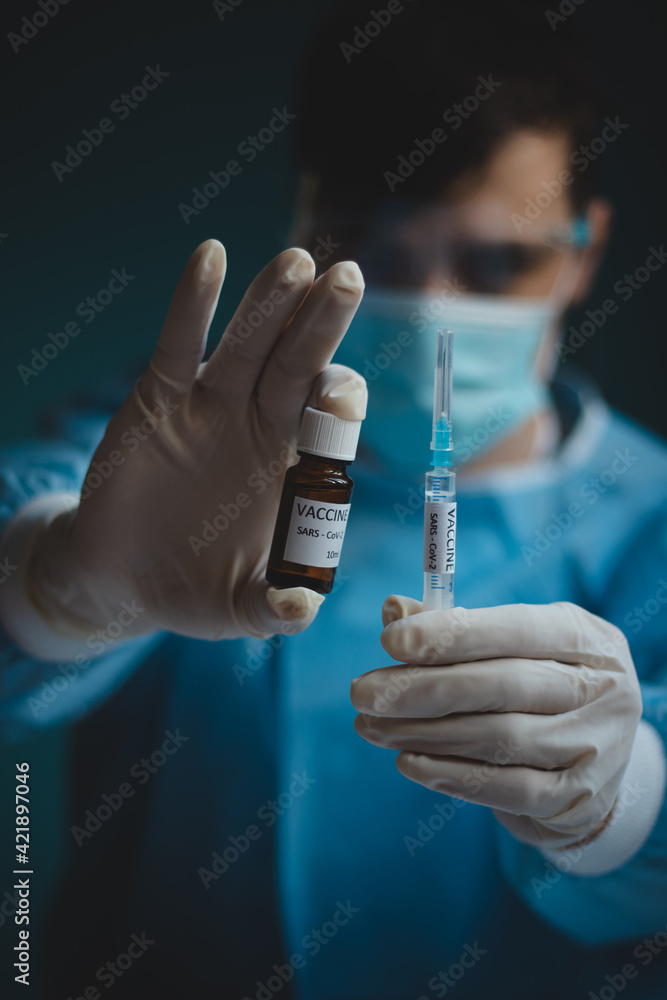doctor in a protective shield, veil, and sterile gloved suit holds a single weapon against the Coronavirus pandemic. A scientist holds an ampoule against a Covid-19 and a syringe needle doctor in a protective shield, veil, and sterile gloved suit holds a single weapon against the Coronavirus pandemic. A scientist holds an ampoule against a Covid-19 and a syringe needle
