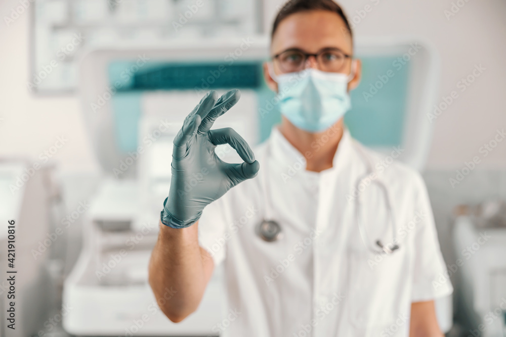 Young doctor with rubber gloves and face mask standing in hospital and showing okay. Corona virus is going down. Young doctor with rubber gloves and face mask standing in hospital and showing okay. Corona virus is going down.