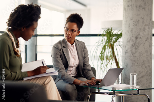Foto African American businesswoman works on laptop while being mentor to a newcomer who is writing notes during the meeting