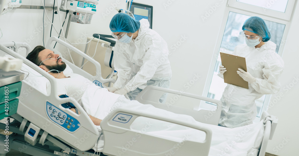 Doctors in protective suits and masks are examining the infected coronavirus young man patient in the hospital ward Doctors in protective suits and masks are examining the infected coronavirus young man patient in the hospital ward