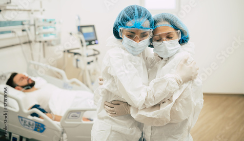 Two confident doctors in protective safety suits standing on background of a patient with coronavirus