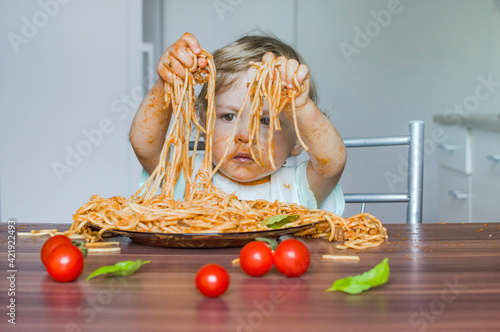 Funny baby child getting messy eating spaghetti with tomato sauce from a large plate, by itself with his hands, at home