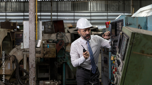 A beard mechanical engineer, supervisor or factory manager is communicating with his staff by using radio or handhold walkie talkie in a warehouse. Smart manager with helmet is checking a mission