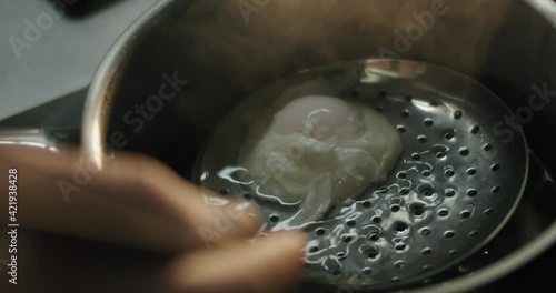 The chef's hand with a slotted spoon takes out a poached egg from boiling water in slow motion