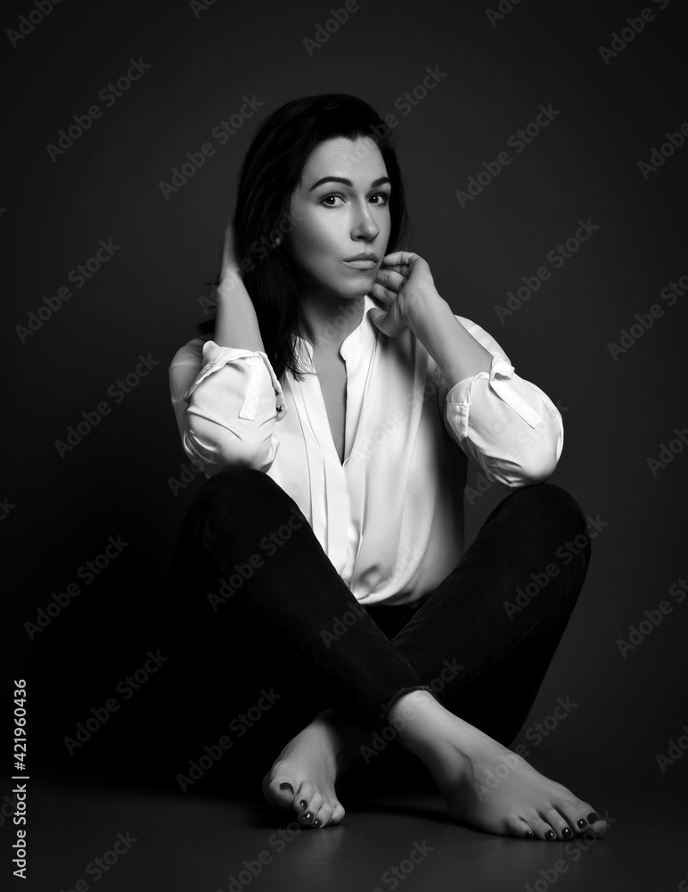 Mature slim good looking brunette woman with perfect skin in white shirt and pants sits barefooted on floor holding hands at hair and cheek. Fashion, stylish casual look for lady concept Mature slim good looking brunette woman with perfect skin in white shirt and pants sits barefooted on floor holding hands at hair and cheek. Fashion, stylish casual look for lady concept