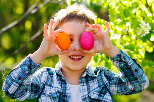 Portrait of a boy having fun in the garden against the background of foliage with Easter eggs held up to his eyes.