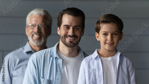 Portrait of smiling three generations of Caucasian men isolated on grey background look at camera. Happy little boy son with young 30s father and older 60s grandfather show family unity and bonding.