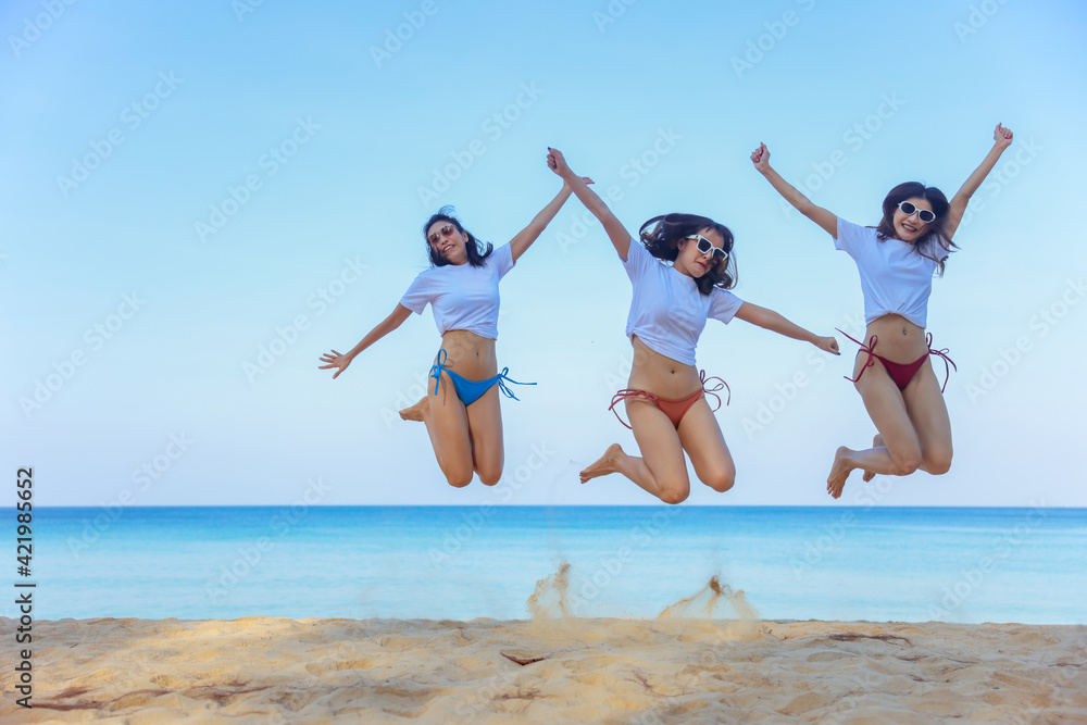 Portrait of three beautiful young asian women friends, wearing bikini and stylish sunglasses jumping on the seashore looking at camera smile. Blue sea and sky in the background Portrait of three beautiful young asian women friends, wearing bikini and stylish sunglasses jumping on the seashore looking at camera smile. Blue sea and sky in the background
