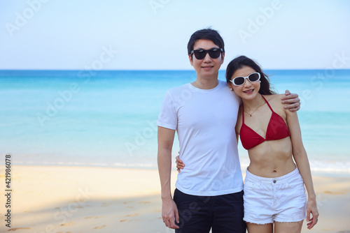 Portrait of beautiful young women and boyfriend asian, wearing rea bikini and stylish white sunglasses standing on the seashore looking at camera smile. Blue sea and sky in the background