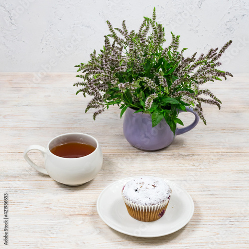 Cupcake on the background of a mug of tea and mint flowers on a white wooden table. Tea party with a cupcake.